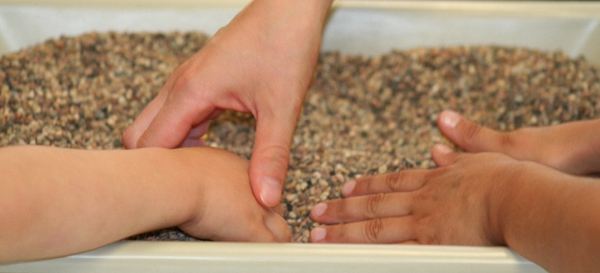Ergotherapie - Kinderhand geführt von anderer Hand im Kiesbett